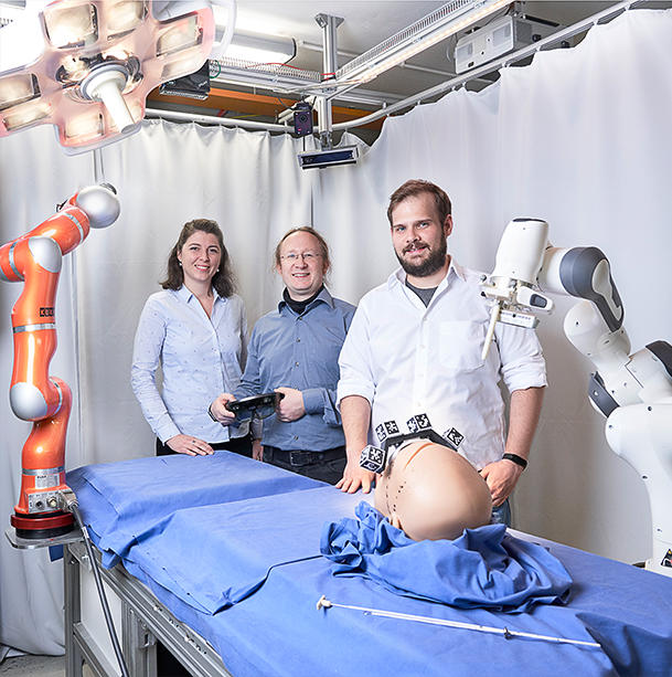 Jun.-Prof. Franziska Mathis-Ullrich, Prof. Björn Hein und Christian Kunz stehen neben Roboter, der Chirurgen bei der Bestimmung von Einstichstelle und -winkel unterstützt.